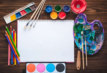 Set of paints, pencils, tools for painting and blank white paper sheet of sketchbook on vintage wooden background. Top view.の写真素材