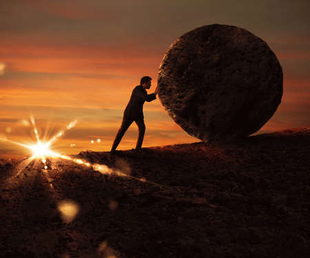 Man pushes a boulder uphill at sunsetの写真素材