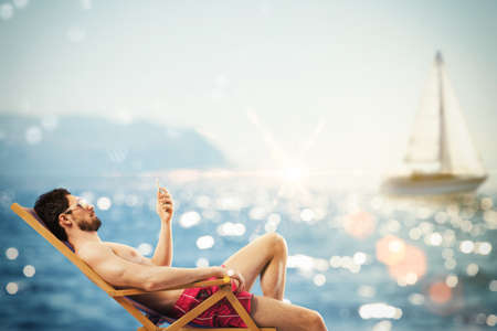 Man with sunglasses and cellphone at sea on deckchairの写真素材