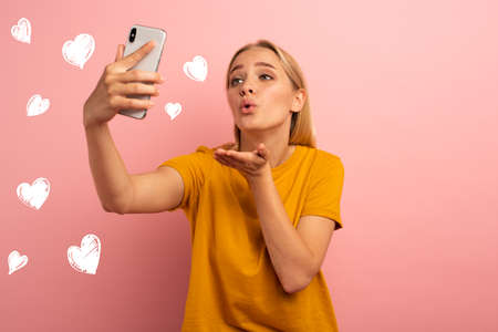 Blonde cute girl sends hearts on her smartphone. Happy and lovely expression face. Pink background