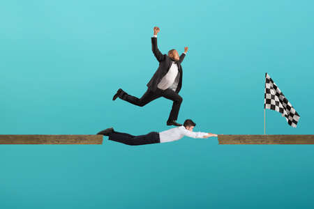 Businessman uses his body like a bridge to help his coullague to reach the flag. concept of teamwork and partnership. Cyan backgroundの写真素材