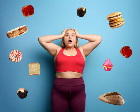 Fat girl in fitness suite wants to start a diet but has doubts about the food to buy. Cyan backgroundの写真素材