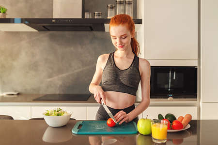 Athletic young red haired woman in the home kitchen prepares a vegetable centrifuged and a saladの写真素材