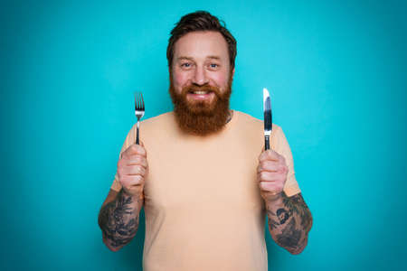 Man with tattoos is ready to eat with cutlery in hand. cyan backgroundの写真素材