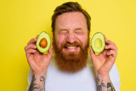 Happy man with beard and tattoos holds an avocadoの写真素材