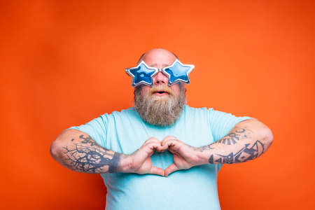 Fat man with beard, tattoos and sunglasses makes heart shape with hands