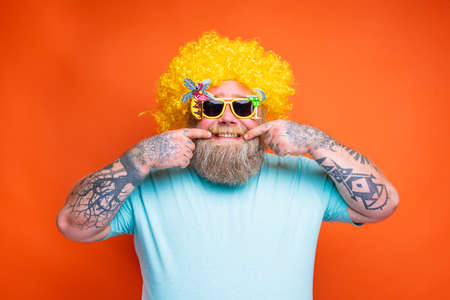 Fat happy man with beard, tattoos and sunglasses has fun with the yellow wig