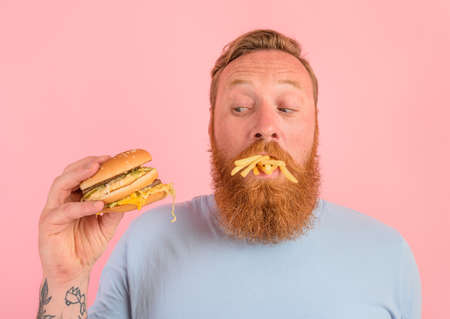 Thoughtful man with beard and tattoos eats a sandwitch with hamburger and potatoesの写真素材