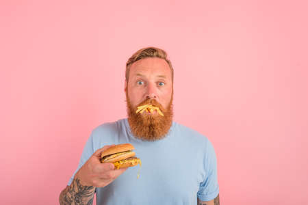 Hungry man with beard and tattoos eats a sandwitch with hamburger and potatoesの写真素材
