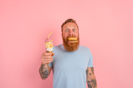 Amazed man with beard and tattoos is undecided if to eat an icecream or a sandwich with hamburgerの写真素材