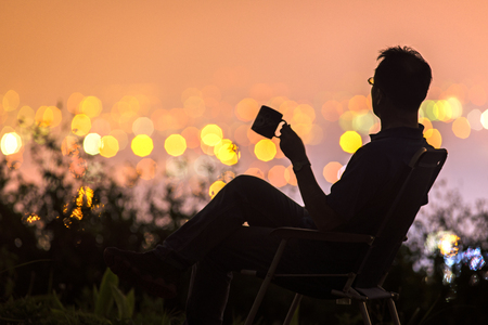 A man holding a cup and sitting on a hill with background of city night light.の写真素材