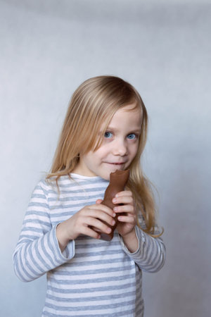 Blonde girl in striped jacket on a light background bites chocolate hare in front of her face smilesの写真素材