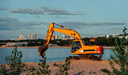 Orange excavator is working at the river bank. Sand, water and trees on background.の写真素材