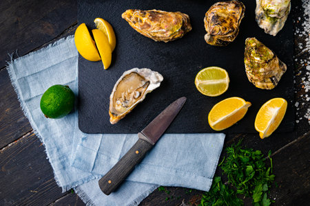 Fresh oysters on a black stone background. Top view. Oyster dinner in restaurant. Dark background. Seafood, Gourmet food. Oyster glove, oyster opening knifeの写真素材
