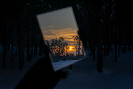 Winter forest at sunset. Reflection of the sun. The hand holds the mirror. Landscape in the mirror.の写真素材