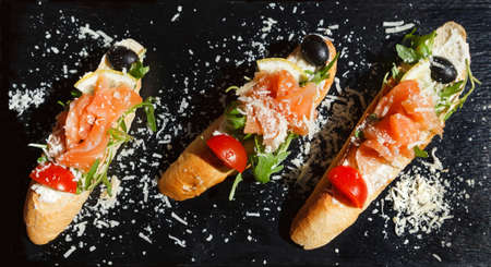 Three salmon sandwiches decorated with herbs, lemon, olives, grated cheese and tomato on a black Board made of natural slate. Flat lay, top viewの写真素材