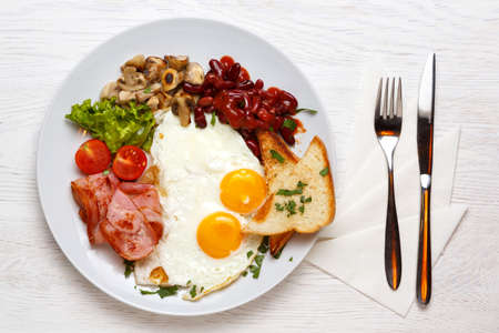 English Breakfast, fried eggs, ham, mushrooms on a white plate with a knife and fork, on a white...