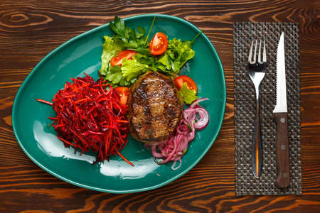 Beef steak with a side dish of carrots, beets and tomatoes on a green plate, on a dark wooden table. Top viewの写真素材