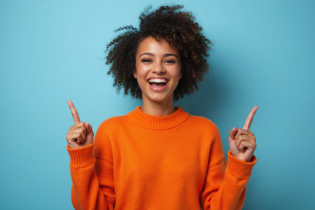 A cheerful woman with curly hair wearing an orange sweater, smiling brightly, and pointing upwards with both hands against a blue background.の素材