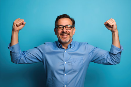 A joyful man with glasses and a blue shirt excitedly raises his fists in triumph against a blue background.の素材