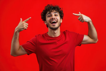 A man with curly hair and a red shirt is pointing upwards with both hands, smiling broadly against a red background.の素材