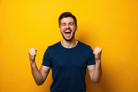 A joyful man with a wide smile and closed eyes, wearing a dark blue t-shirt, celebrating with raised fists against a bright yellow background.の素材