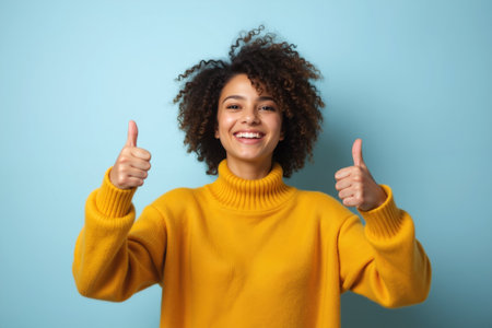 A cheerful woman with curly hair wearing a yellow sweater is smiling and giving two thumbs up against a blue background.の素材