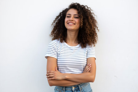 A beautiful woman with curly hair smiling, wearing a striped shirt, standing against a white wall.の素材