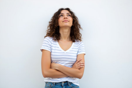 A young woman with curly hair and a striped t-shirt stands confidently looking up with arms crossed against a plain white wall.の素材
