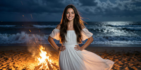 Smiling young woman in flowing white dress posing on windy beach with burning bonfire, dramatic sea, and dark cloudy sky.の素材