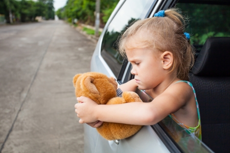 sad little girl sitting near the window in the car with toy in her handsの写真素材
