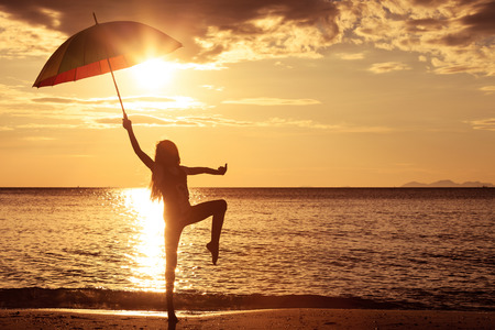 Happy girl with an umbrella jumping on the beach at the sunset timeの写真素材