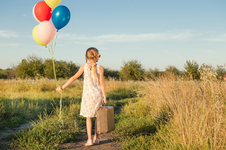 Happy little girl playing on road at the day time. She standing in the field and holding in hands suitcase and balloons. Kid having fun on the nature. Concept of happiness.の写真素材