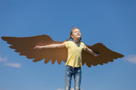 Little girl playing with cardboard toy wings in the park at the day time. Concept of happy game. Child having fun outdoors. Picture made on the background of blue sky.の写真素材
