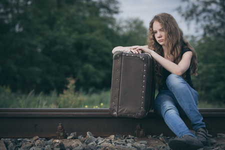 Portrait of young sad ten girl sitting with suitcase outdoors  on the railway at the day time. Concept of sorrow.の写真素材