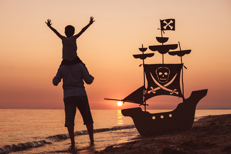 Father and son playing on the beach at the sunset time. They playing with a cardboard pirate ship. People having fun outdoors. Concept of summer vacation and friendly family.の写真素材
