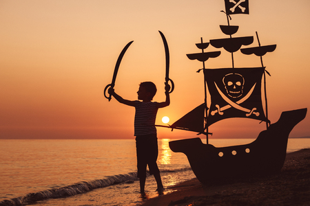 One happy little boy standing on the beach at the sunset time. He playing with a cardboard pirate ship.  Kid having fun outdoors. Concept of summer vacation.の写真素材