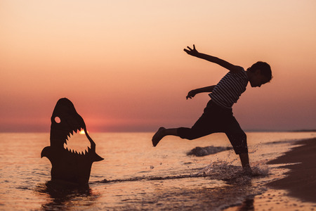 One happy little boy running on the beach at the sunset time. He playing with a cardboard shark.  Kid having fun outdoors. Concept of summer vacation.の写真素材
