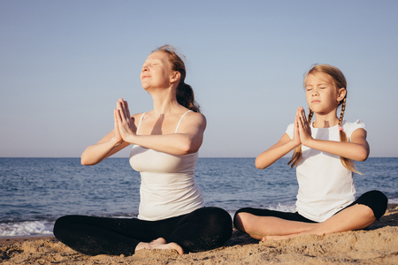 Mother and daughter doing yoga exercises  on the beach at the day time. People having fun outdoors. Concept of summer vacation and friendly family.の写真素材