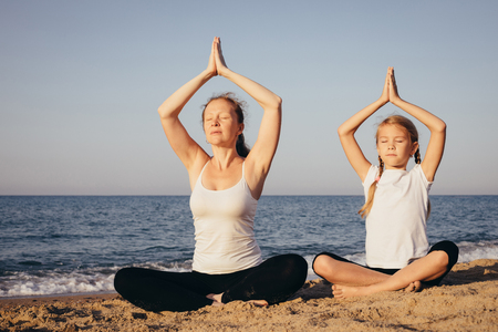 Mother and daughter doing yoga exercises  on the beach at the day time. People having fun outdoors. Concept of summer vacation and friendly family.の写真素材