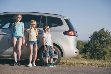 Happy brother and his two sisters are standing near the car at the day time. Children having fun outdoors. Concept of the family is ready for travel.の写真素材