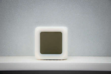 Conceptual blurred closeup of square shape empty white cube clock standing on office shelf on grey...