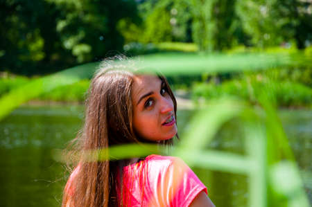 young sexy smiling woman or girl with pretty cute face and long brunette hair in pink shirt has braces or brackets on teeth near river or sea water sunny outdoor on natural backgroundの写真素材