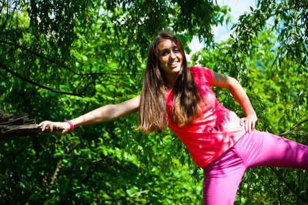 young sexy smiling woman or girl with pretty cute face and long brunette hair in pink shirt posing on tree trunk with green leaves sunny outdoor on natural backgroundの写真素材