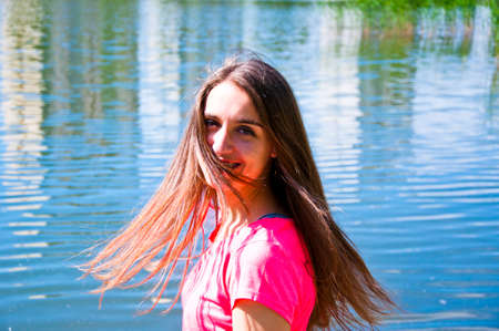 young sexy smiling woman or girl with pretty cute face and long brunette hair in pink shirt near river or sea water sunny outdoor on natural backgroundの写真素材
