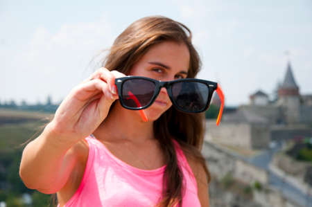 young woman or girl with long brunette hair and pretty face in pink summer vest sunny day outdoor on beautiful castle or palace background holding sunglasses or glasses in hand, selective focusの写真素材
