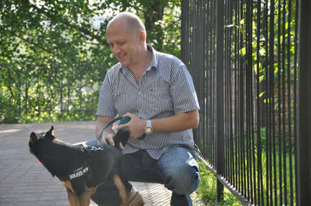 handsome bald man with smiling face near iron fence with beautiful cute black and brown dog pet friend outdoor in summer on green sunny dayの写真素材