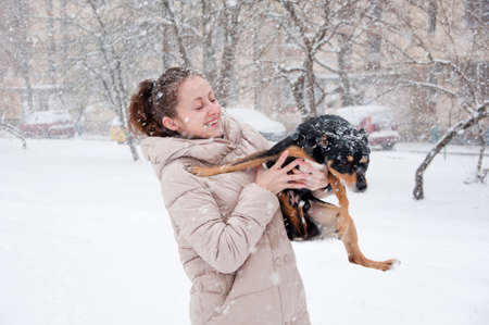 pretty cute young smiling girl or sexy woman in beige winter coat snowy cold winter on natural white background stands in snow and snowflakes with black dog or puppy petの写真素材