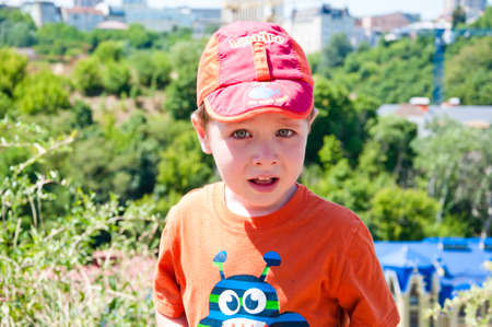 cute small baby boy or child with crying face in red cap and orange shirt sunny outdoor on natural green trees background in summerの写真素材