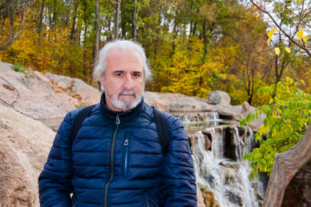 mature man with graying hair and beard mustache at waterfall with autumn forest, copy space, nature.